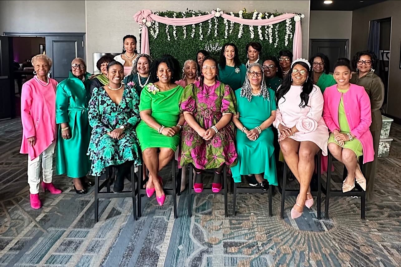 Iota Zeta Omega sorors in pink and green gathered in front of a greenery wall